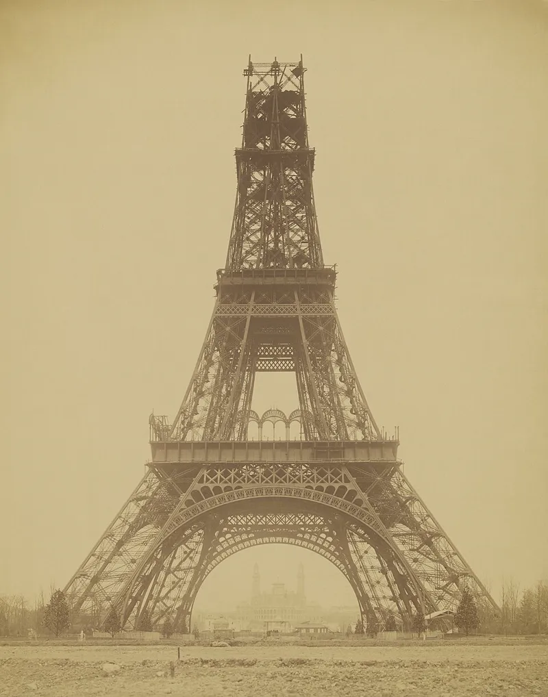 Sepia photograph of the Eiffel Tower under construction, finished up to the second level, with scaffolding at the top